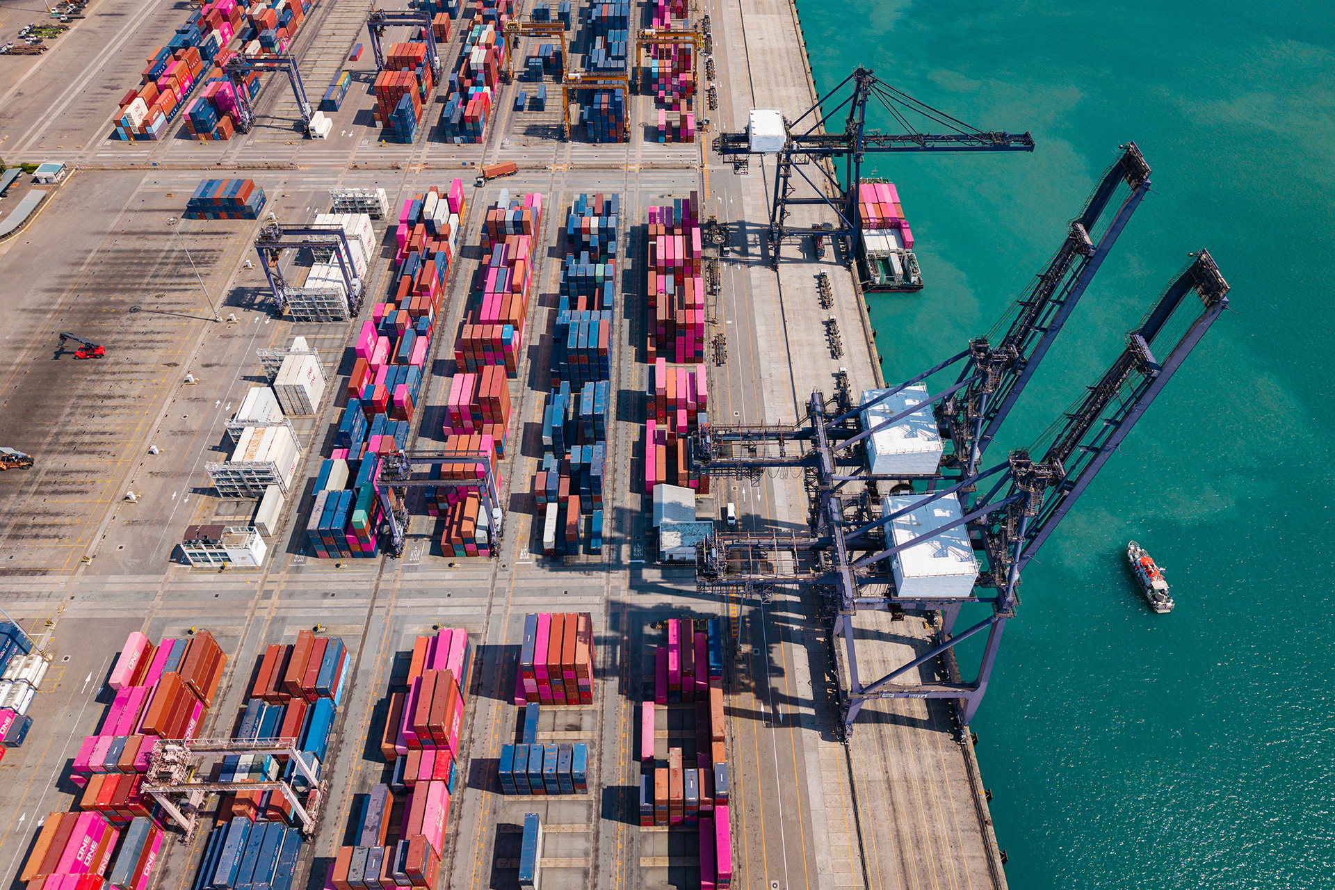 Aerial view of busy container terminal and cargo cranes Drone shot of a bustling shipping port with colorful containers and giant cargo cranes beside bright turquoise ocean waters.