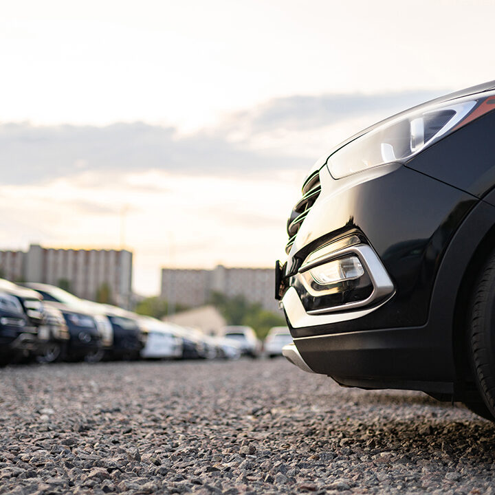 A row of cars parked outside on a gravel lot, featuring a close-up of a black vehicle's front.